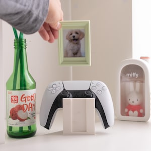 May include: A green glass bottle with a "Good Day" label, a white and black game controller, a small framed photo of a dog, and a Miffy figurine in a clear case. The photo is being held by a person.