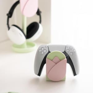 May include: A light gray game controller rests on a green and pink tulip-shaped stand. A matching stand holds white and black headphones. The stands and headphones are on a white surface, with a green leaf-shaped object in the foreground.