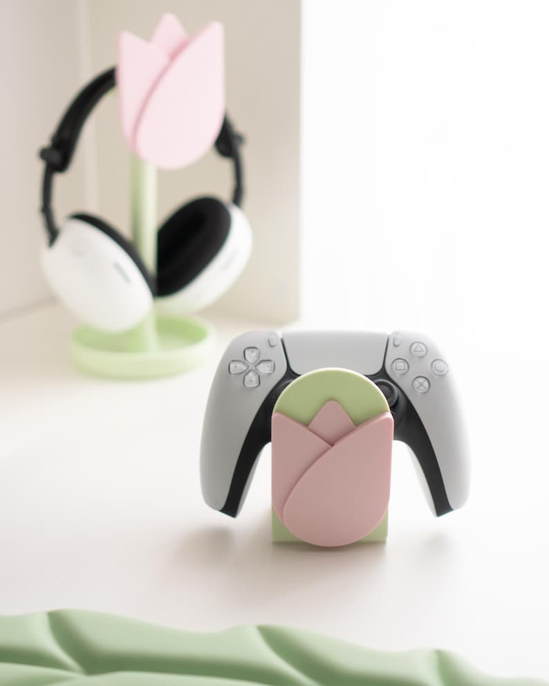 May include: A light gray game controller rests on a green and pink tulip-shaped stand. A matching stand holds white and black headphones. The stands and headphones are on a white surface, with a green leaf-shaped object in the foreground.