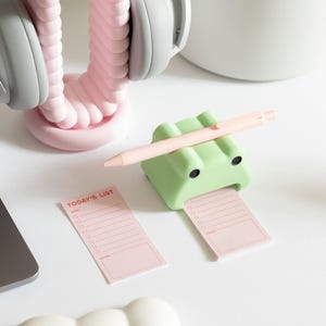 May include: A green frog-shaped sticky note holder with a pink pen resting on top. The holder is holding a pink sticky note with the text "TODAY'S LIST" in red. There is a white cloud-shaped object in the lower left corner of the image.