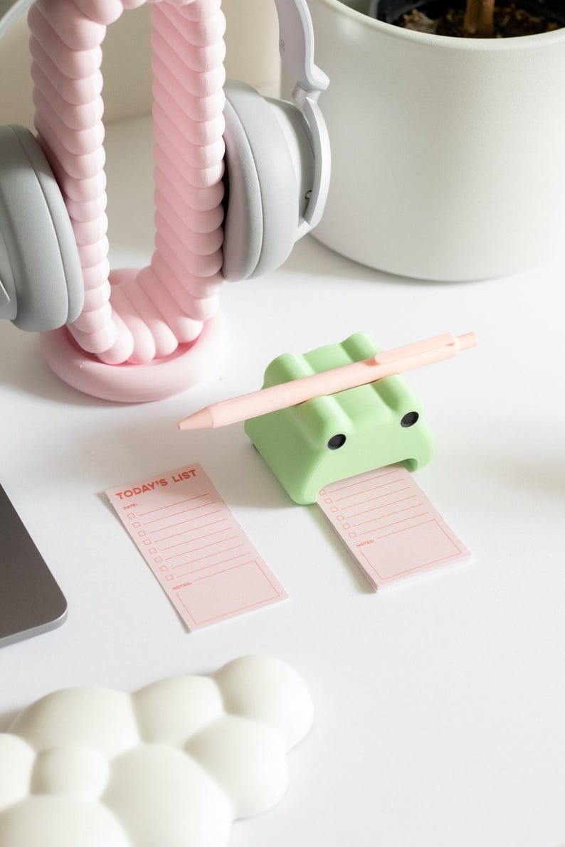 May include: A green frog-shaped sticky note holder with a pink pen resting on top. The holder is holding a pink sticky note with the text "TODAY'S LIST" in red. There is a white cloud-shaped object in the lower left corner of the image.