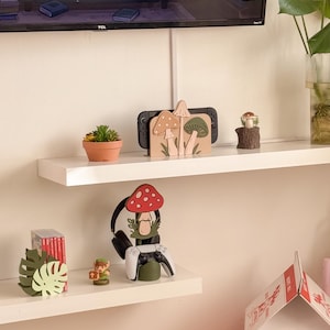 Pu&ograve; includere: Un'esposizione decorativa su scaffale con una Nintendo Switch, un supporto per cuffie e varie decorazioni a tema funghi. Il supporto per cuffie ha una parte superiore a forma di fungo rosso con pois bianchi. Sono presenti anche una piccola pianta grassa in vaso e un fungo in miniatura su un ceppo.