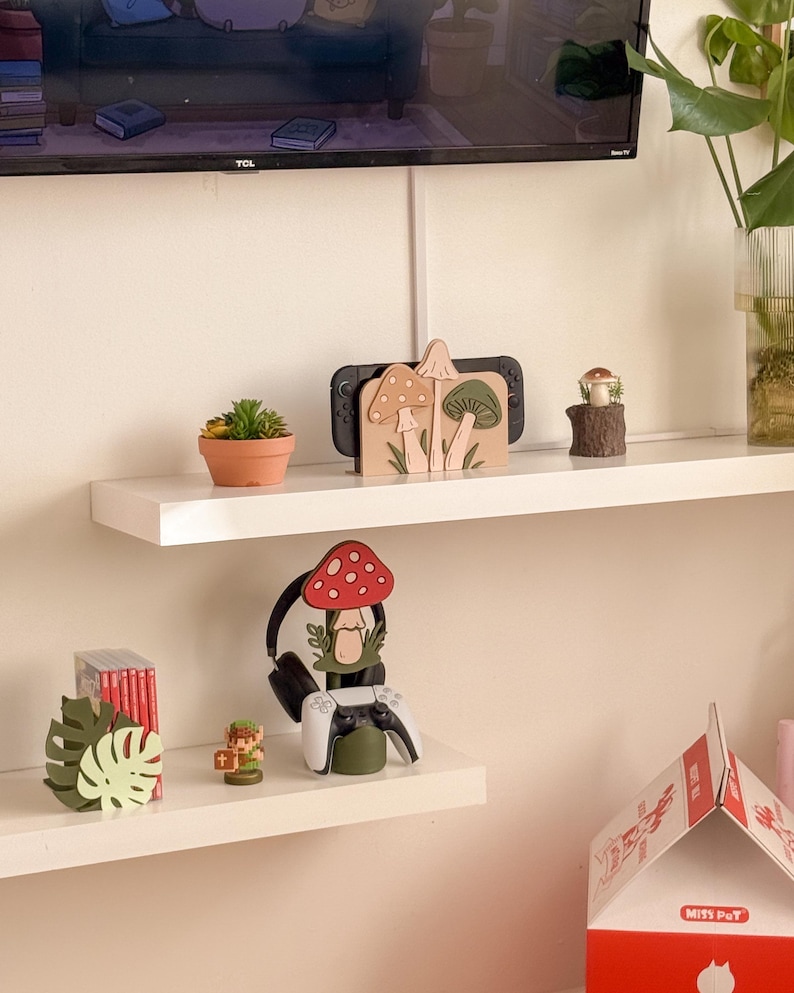 Pu&ograve; includere: Un'esposizione decorativa su scaffale con una Nintendo Switch, un supporto per cuffie e varie decorazioni a tema funghi. Il supporto per cuffie ha una parte superiore a forma di fungo rosso con pois bianchi. Sono presenti anche una piccola pianta grassa in vaso e un fungo in miniatura su un ceppo.