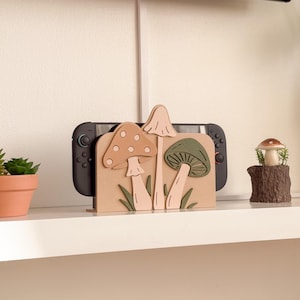 Pu&ograve; includere: Un supporto per console di videogiochi in legno con un design a fungo nei toni del beige, rosa e verde. Il supporto contiene una console di gioco nera. Sono visibili anche una piccola pianta in vaso e una statuina a forma di fungo.