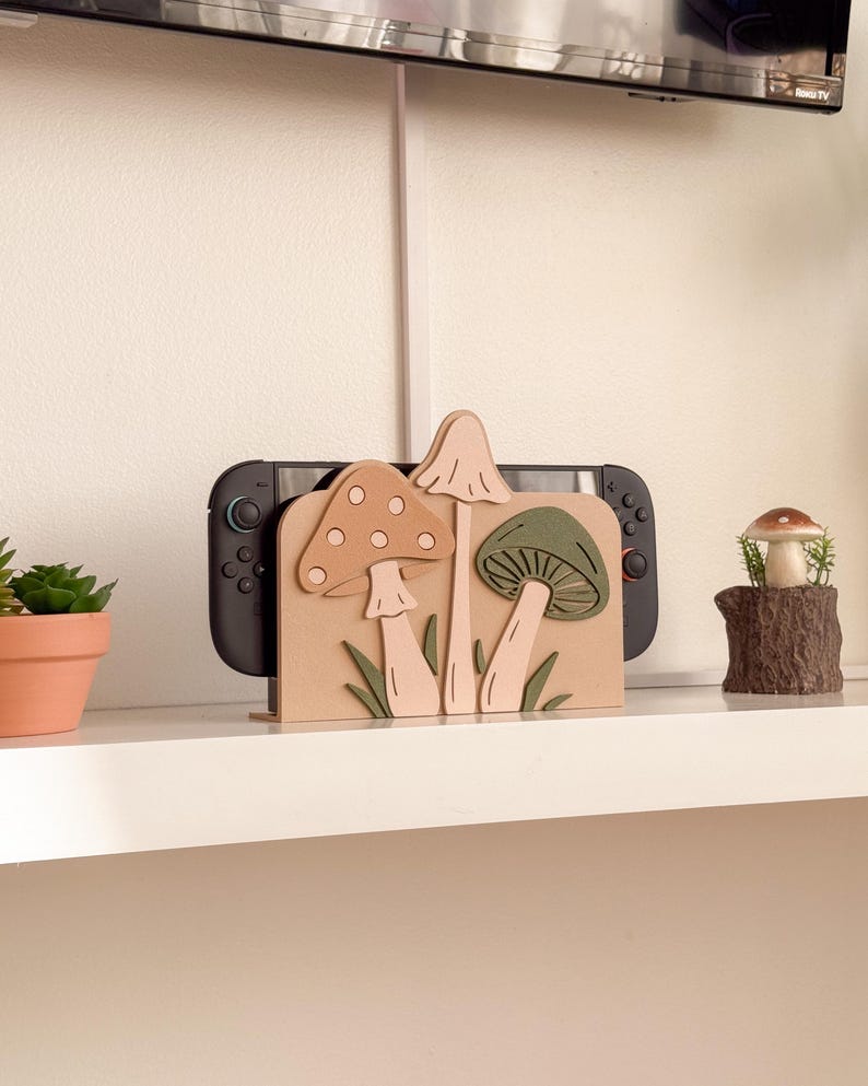 Pu&ograve; includere: Un supporto per console di videogiochi in legno con un design a fungo nei toni del beige, rosa e verde. Il supporto contiene una console di gioco nera. Sono visibili anche una piccola pianta in vaso e una statuina a forma di fungo.