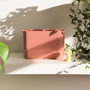 May include: A textured, rectangular, coral-colored storage container with a stepped design. The container sits on a white shelf, with a small yellow figurine and trailing green plants in the background. The container is empty and ready to be filled.