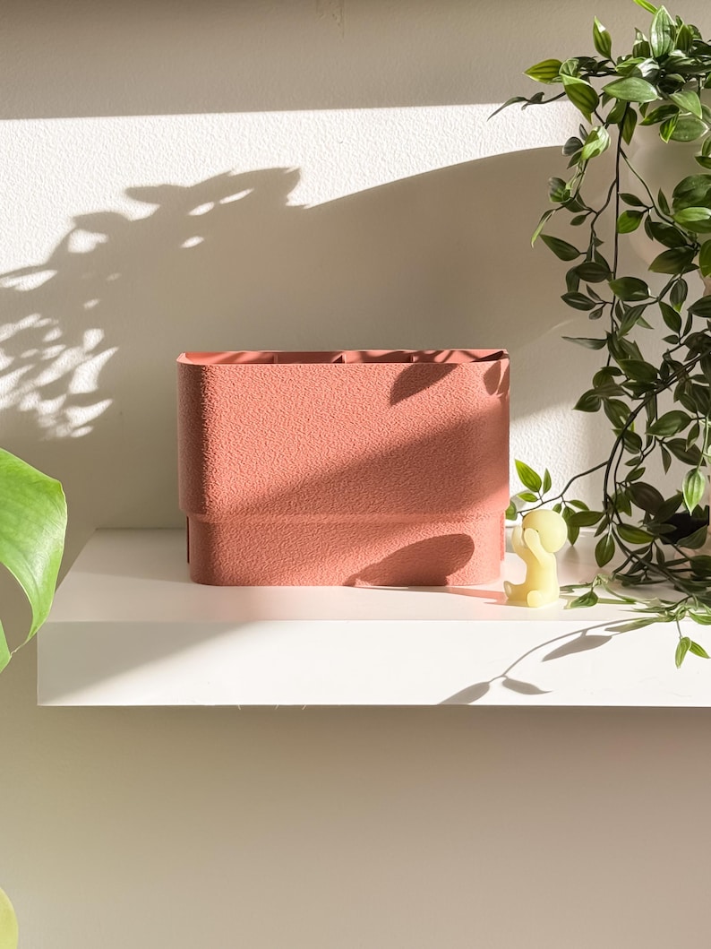 May include: A textured, rectangular, coral-colored storage container with a stepped design. The container sits on a white shelf, with a small yellow figurine and trailing green plants in the background. The container is empty and ready to be filled.