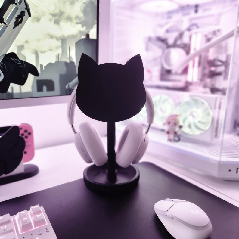 Puede incluir: Un soporte negro para auriculares con forma de gato sostiene unos auriculares blancos. El soporte est&aacute; sobre una alfombrilla de escritorio negra, con un teclado y un rat&oacute;n blancos. Un monitor de ordenador y una consola de juegos son visibles en el fondo.
