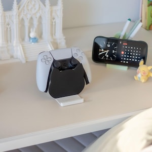 May include: A black cat-shaped controller stand holding a white and black gaming controller. The stand features a flat base and a silhouette of a cat head. The controller is positioned upright in the center of the stand, showcasing its buttons and joysticks clearly. The design is simple and functional, providing a stable mount for the gaming device while maintaining a clean look on the desk surface.