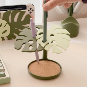 Pu&ograve; includere: Un portapenne verde a forma di foglia di monstera con base in sughero. Il supporto ha uno stelo verde scuro e foglie verde chiaro. Una penna viola e una penna verde sono nel supporto. Una console Switch bianca e verde &egrave; sullo sfondo.