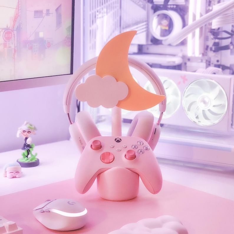 May include: A pink gaming headset stand with a moon and cloud design. The stand holds a pink Xbox controller and a pair of pink headphones.