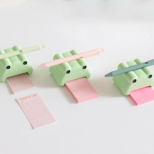 May include: Three green frog-shaped sticky note holders with pens on top. Each frog has a pink sticky note attached to its mouth. One sticky note has the text "TODAY'S LIST" written on it.