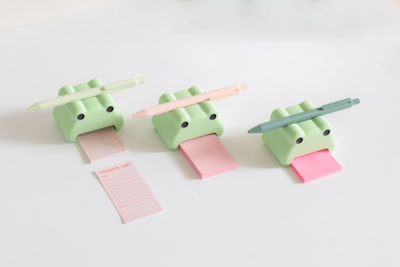 May include: Three green frog-shaped sticky note holders with pens on top. Each frog has a pink sticky note attached to its mouth. One sticky note has the text "TODAY'S LIST" written on it.