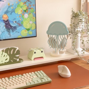 May include: A desk setup featuring a light green keyboard, a white ergonomic mouse, and a light blue jellyfish-shaped headphone stand. Other items include a green frog-shaped desk organizer and a decorative monstera leaf. A computer monitor displays a colorful animated scene.