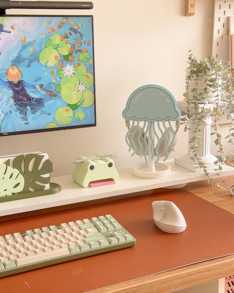 May include: A desk setup featuring a light green keyboard, a white ergonomic mouse, and a light blue jellyfish-shaped headphone stand. Other items include a green frog-shaped desk organizer and a decorative monstera leaf. A computer monitor displays a colorful animated scene.