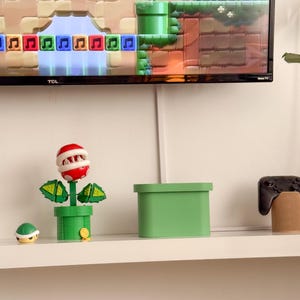 May include: A green plant with a red mouth and teeth, a green pot, and a green box on a white shelf. There is a small green turtle figurine and a black video game controller on the shelf. The TV screen in the background shows a video game with a green pipe and a red mushroom.