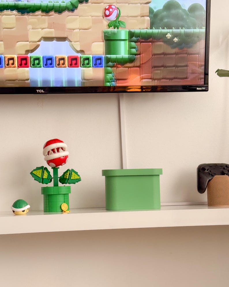 May include: A green plant with a red mouth and teeth, a green pot, and a green box on a white shelf. There is a small green turtle figurine and a black video game controller on the shelf. The TV screen in the background shows a video game with a green pipe and a red mushroom.