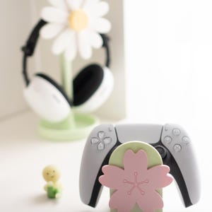 May include: A white and gray game controller rests on a light green and pink flower-shaped stand. A white daisy-shaped headphone stand is in the background. A small yellow figurine is also present.