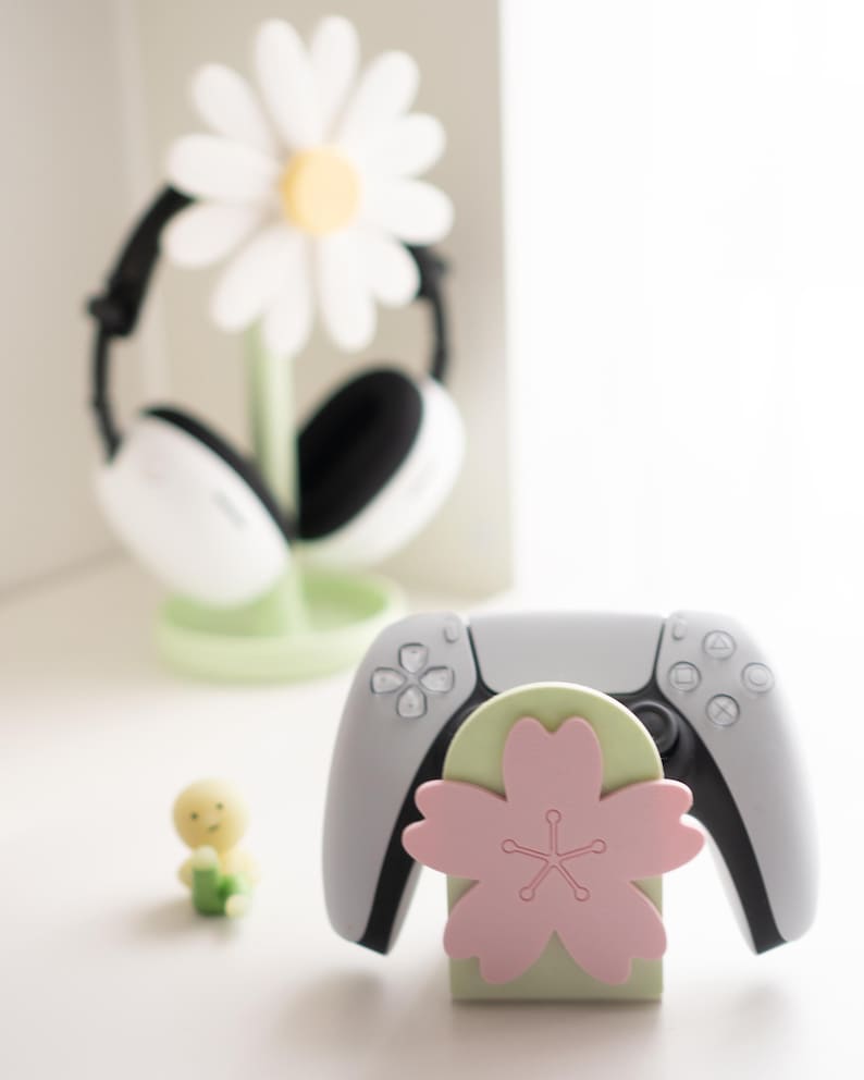 May include: A white and gray game controller rests on a light green and pink flower-shaped stand. A white daisy-shaped headphone stand is in the background. A small yellow figurine is also present.