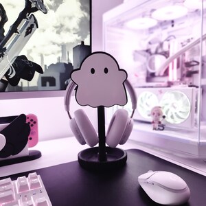 May include: A white ghost-shaped headphone stand with a black base, holding a pair of white headphones. The stand is on a white desk with a white keyboard and a white mouse.
