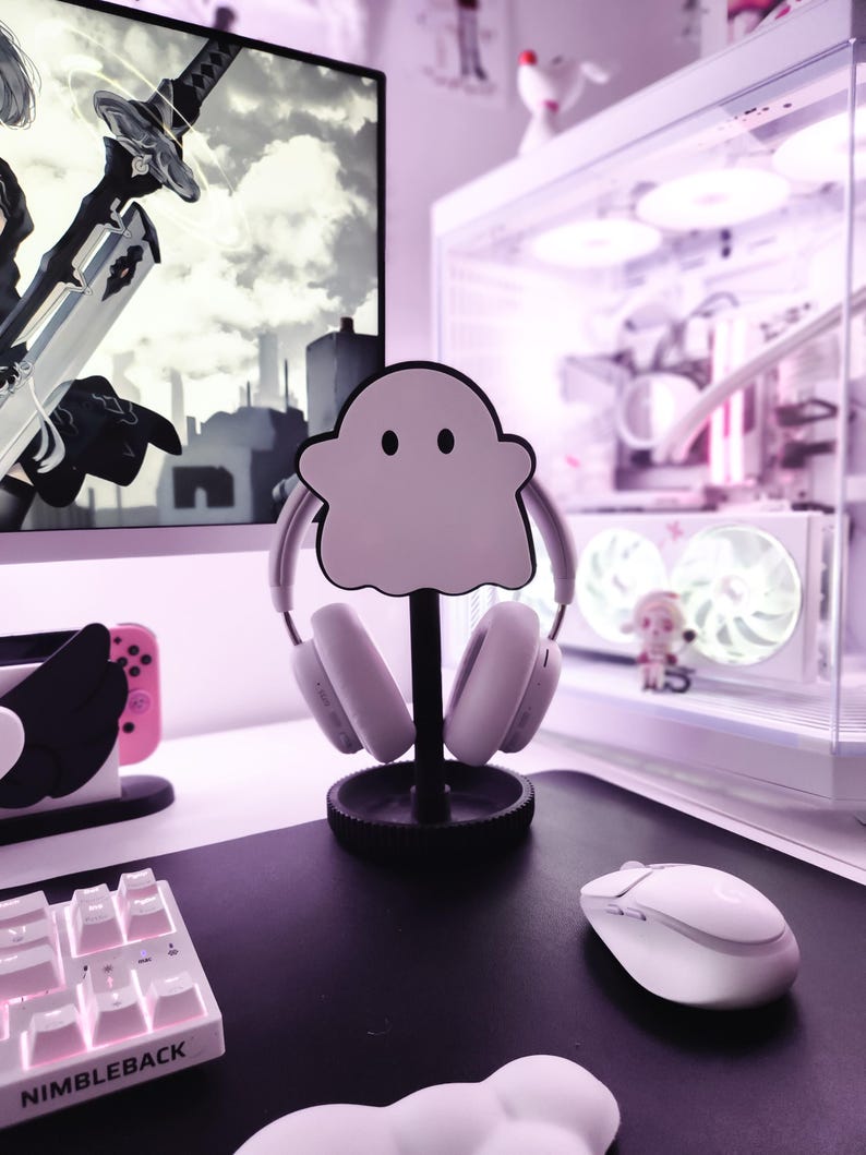 May include: A white ghost-shaped headphone stand with a black base, holding a pair of white headphones. The stand is on a white desk with a white keyboard and a white mouse.