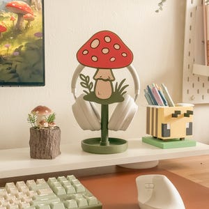 Pu&ograve; includere: Un'impostazione da scrivania stravagante con un supporto per cuffie a forma di fungo con cappello rosso e pois bianchi. Sono visibili anche una piccola fioriera a fungo e un portapenne a tema Minecraft, insieme a una tastiera e un mouse.