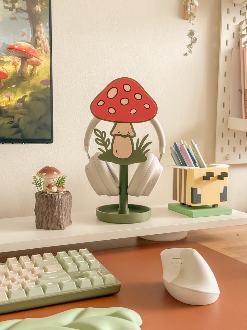 Pu&ograve; includere: Un'impostazione da scrivania stravagante con un supporto per cuffie a forma di fungo con cappello rosso e pois bianchi. Sono visibili anche una piccola fioriera a fungo e un portapenne a tema Minecraft, insieme a una tastiera e un mouse.