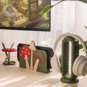 Pu&ograve; includere: Un allestimento da scrivania con un supporto per console di gioco a tema funghi verdi e un supporto per cuffie abbinato. Il supporto mostra una console di gioco nera. Le cuffie bianche poggiano sul supporto. Un monitor mostra un'illustrazione accogliente.