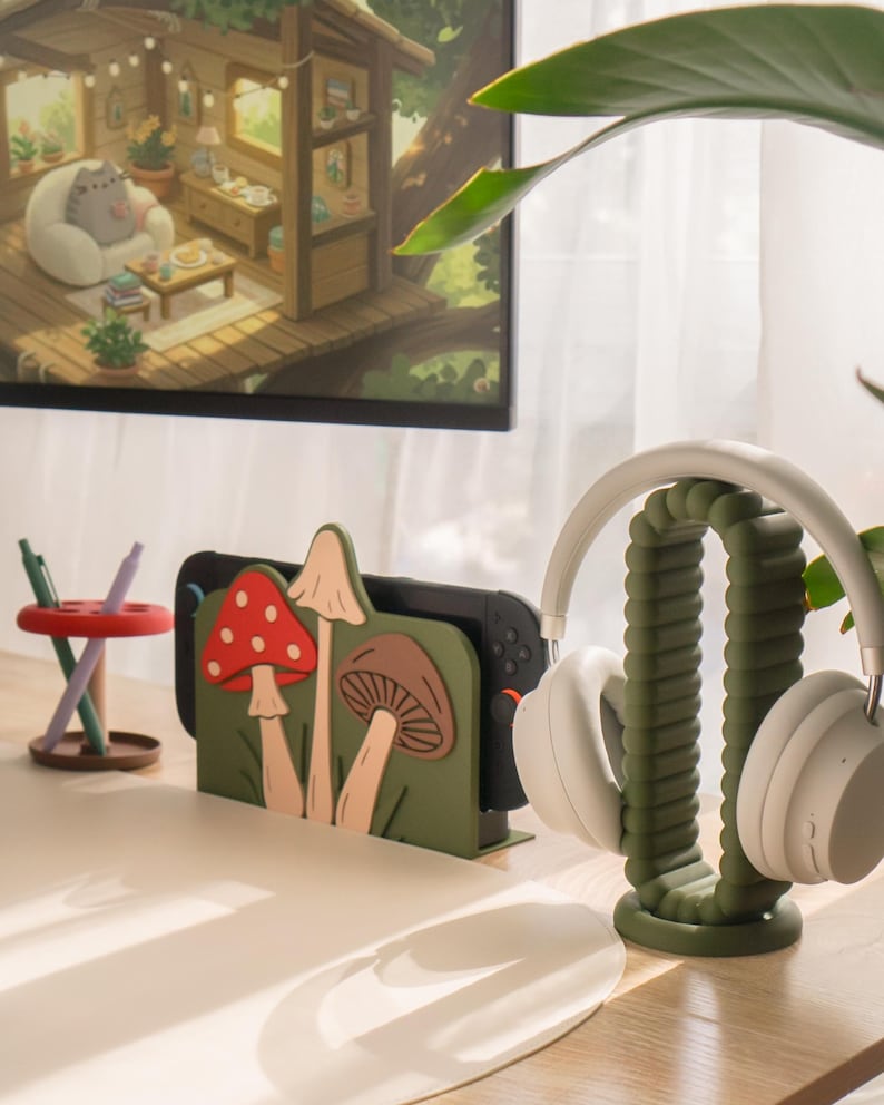 Pu&ograve; includere: Un allestimento da scrivania con un supporto per console di gioco a tema funghi verdi e un supporto per cuffie abbinato. Il supporto mostra una console di gioco nera. Le cuffie bianche poggiano sul supporto. Un monitor mostra un'illustrazione accogliente.