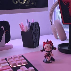 May include: A black coffin-shaped pen holder with ornate detailing, filled with various pens and markers. A small figurine and a keyboard with pink keycaps are also visible. The scene is set on a pink surface.