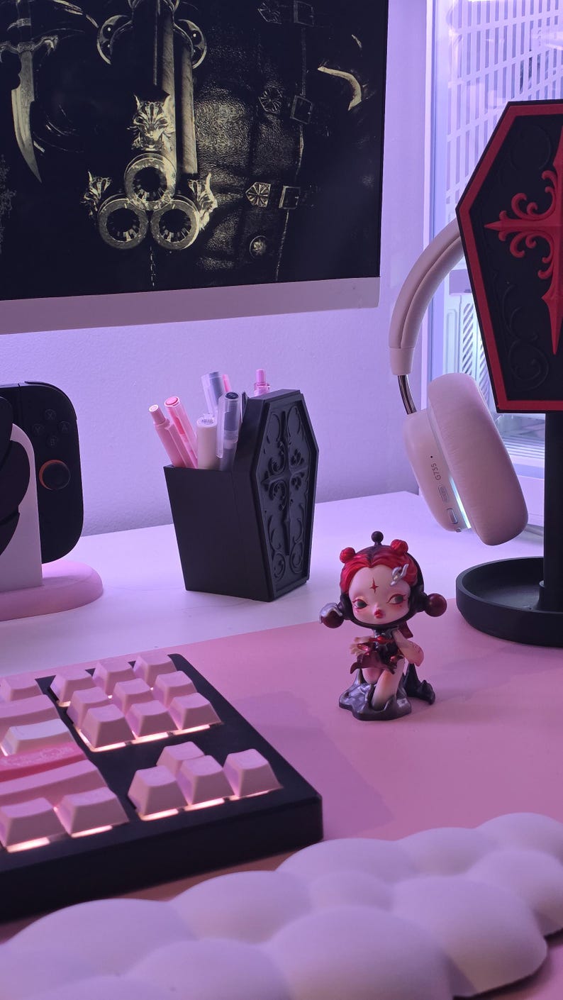 May include: A black coffin-shaped pen holder with ornate detailing, filled with various pens and markers. A small figurine and a keyboard with pink keycaps are also visible. The scene is set on a pink surface.