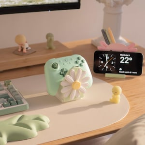 May include: A light green video game controller stand featuring a large white daisy flower with a yellow centre on the front. The stand is designed to hold a controller upright and is positioned on a desk with other small desk accessories.