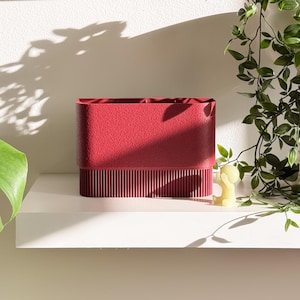 May include: A textured, rectangular, red container with a ribbed lower section sits on a white shelf. A small yellow figurine and trailing green plants are to the right. The container is likely for organizing items.