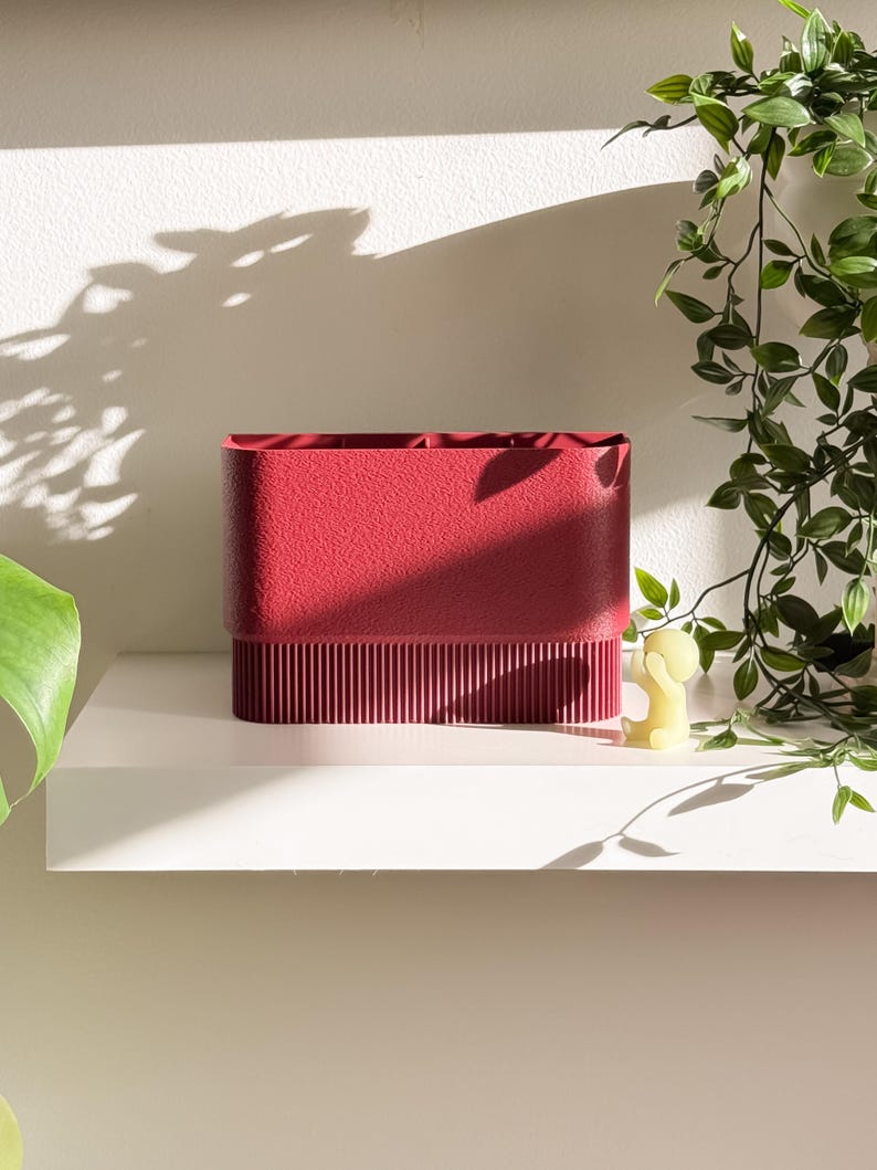 May include: A textured, rectangular, red container with a ribbed lower section sits on a white shelf. A small yellow figurine and trailing green plants are to the right. The container is likely for organizing items.