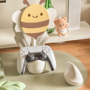 May include: A gaming setup featuring a white and gray PlayStation 5 controller resting in a white holder. A cute bee-shaped headphone stand holds a pair of headphones. A small teddy bear figurine and a white ergonomic mouse are also visible.