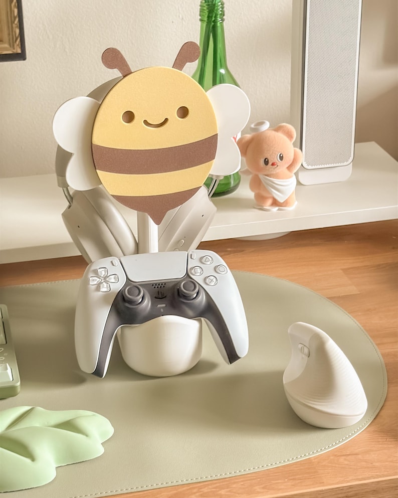 May include: A gaming setup featuring a white and gray PlayStation 5 controller resting in a white holder. A cute bee-shaped headphone stand holds a pair of headphones. A small teddy bear figurine and a white ergonomic mouse are also visible.
