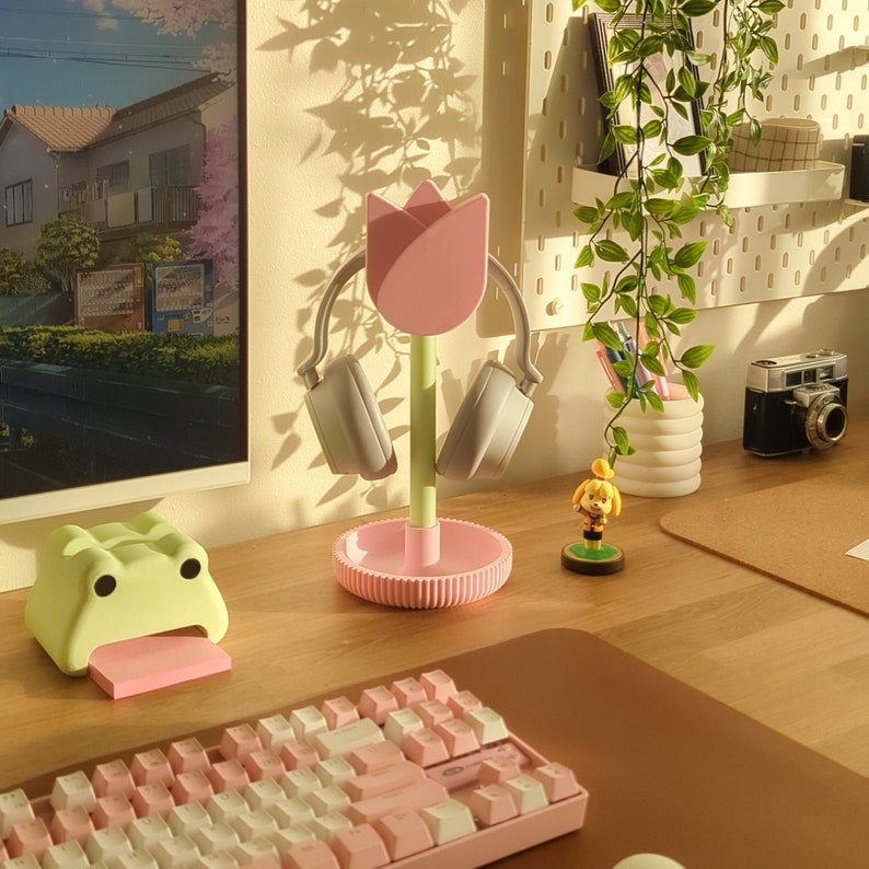 Puede incluir: Un soporte para auriculares en forma de tulip&aacute;n rosa con un tallo verde y unos auriculares grises. El soporte est&aacute; sobre un escritorio de madera con un teclado, un porta notas adhesivas en forma de rana verde y una peque&ntilde;a figura.