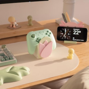 May include: A light green gaming controller rests on a white stand featuring a pink tulip design. Nearby, a pink axolotl-shaped holder supports a smartphone displaying a clock and weather. A small yellow figurine sits on the desk next to a leaf-shaped mat.