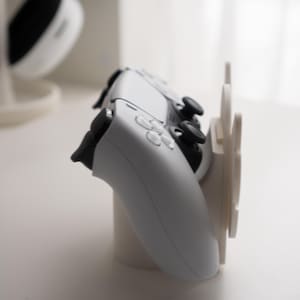 May include: A white gaming controller rests on a white stand, with a matching headset in the background. The controller has black buttons and joysticks. The stand has a circular base and a vertical support.