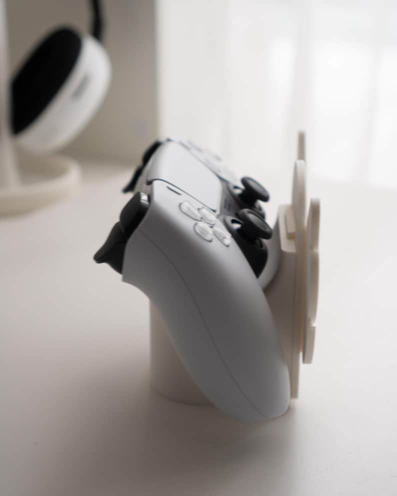 May include: A white gaming controller rests on a white stand, with a matching headset in the background. The controller has black buttons and joysticks. The stand has a circular base and a vertical support.