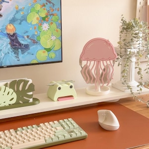 May include: A desk setup featuring a pink jellyfish-shaped headphone stand, a green frog-shaped pen holder, and a mint-colored keyboard. A white computer mouse sits on a brown desk mat. A computer monitor displays a colorful scene.