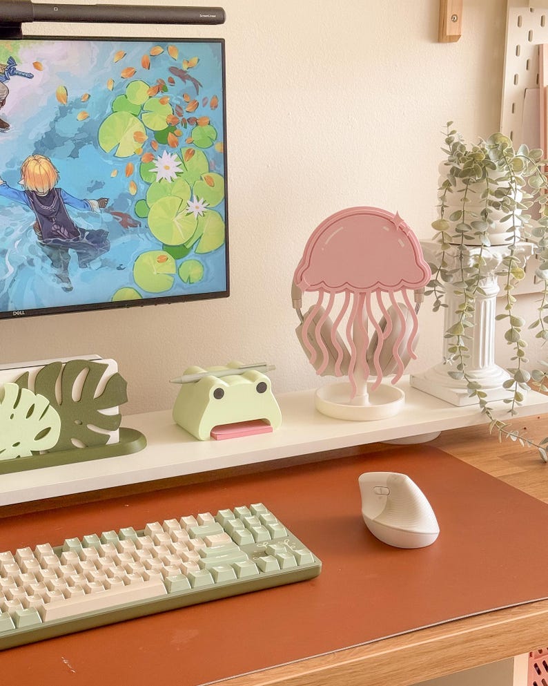 May include: A desk setup featuring a pink jellyfish-shaped headphone stand, a green frog-shaped pen holder, and a mint-colored keyboard. A white computer mouse sits on a brown desk mat. A computer monitor displays a colorful scene.
