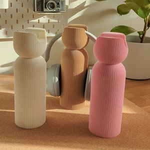 May include: Three decorative bottles with a ribbed design. The bottles are white, beige, and pink. The bottles are sitting on a cork surface.