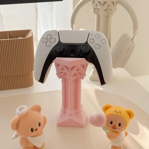 May include: A white and black video game controller rests on a pink decorative stand. A pair of white headphones and two small figurines, one a bear and the other a baby, are also present. The figurines are holding a heart-shaped cotton candy.