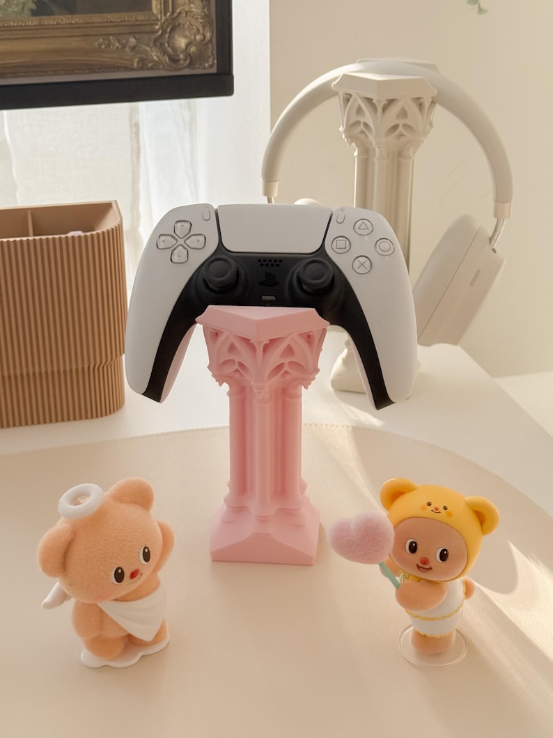 May include: A white and black video game controller rests on a pink decorative stand. A pair of white headphones and two small figurines, one a bear and the other a baby, are also present. The figurines are holding a heart-shaped cotton candy.