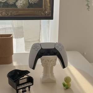 May include: A white and black video game controller rests on a decorative white stand. A miniature black grand piano and a small yellow and green figurine are also on display. A painting with a gold frame hangs in the background.
