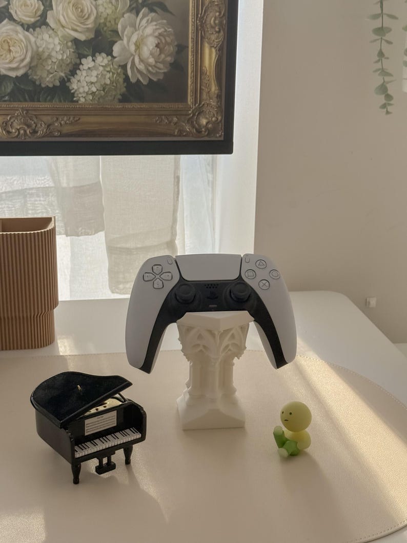 May include: A white and black video game controller rests on a decorative white stand. A miniature black grand piano and a small yellow and green figurine are also on display. A painting with a gold frame hangs in the background.