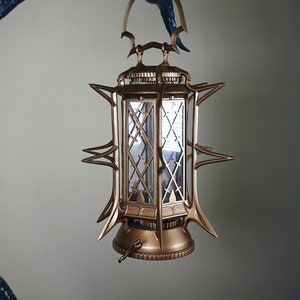 Könnte beinhalten: Eine dekorative Laterne mit goldener Oberfläche, die an einer dunkelblauen, astähnlichen Struktur aufgehängt ist. Die Laterne hat ein geometrisches Design und ist beleuchtet, wodurch eine warme Atmosphäre entsteht. Das Gesamtdesign erinnert an den gotischen Stil.