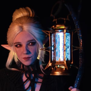 Puede incluir: Una persona con un disfraz de fantasía con orejas puntiagudas y una peluca rubia, sosteniendo una linterna brillante. El disfraz incluye una parte superior negra con flecos y una gargantilla oscura. La linterna tiene un marco dorado y luz azul.