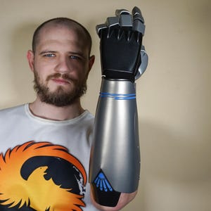 Könnte beinhalten: Ein silberner und schwarzer Prothesenarm mit detaillierter Hand. Der Arm hat ein blaues Akzentband und ein dreieckiges Design in der Nähe des Handgelenks. Die Person trägt ein weißes T-Shirt mit einem großen orange-gelben Aufdruck.